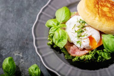 İngiliz çörekleri ve koyu dokular üzerine taze fesleğenle servis edilen pişmiş yumurtalı Benedict. Boşlukları kopyala