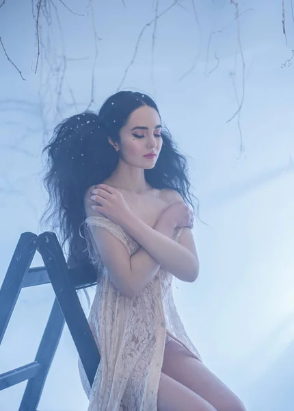 Fairy-tale princess in a peach vintage dress. A girl with an unusual, voluminous hairstyle for long hair with a strong fleece poses on a wooden staircase, in a white room bathed in light and smoke.