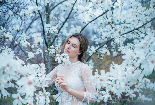 A girl with fair hair, in a transparent dressing-gown posing against a background of blooming, white magnolia. Spring, gentle portraits, as much as possible natural with minimal processing