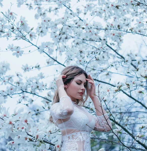 A girl with fair hair, in a transparent dressing-gown posing against a background of blooming, white magnolia. Spring, gentle portraits, as much as possible natural with minimal processing
