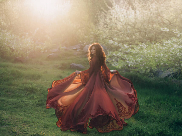 A brunette girl with wavy, thick hair runs to meet the sun at sunset. On the princess is a luxurious, red dress with a long train that flutters in the wind. Background nature early spring. Art photo.