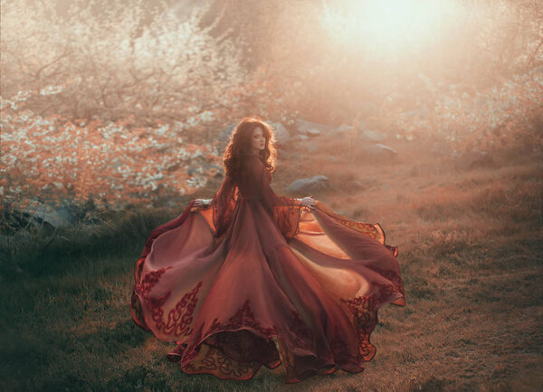 A brunette girl with wavy, thick hair runs to the sun and looks back. The princess has a luxurious, chiffon, red dress that flutters in the wind. Background of a fiery sunset and wild nature.