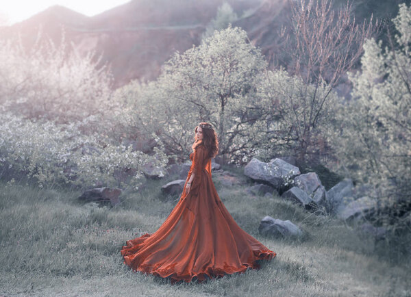 A brunette girl with a golden crown and in a red dress in a long train walking at sunset. Background wildlife, flowering trees, sunlight, lush grass. Artistic photo.