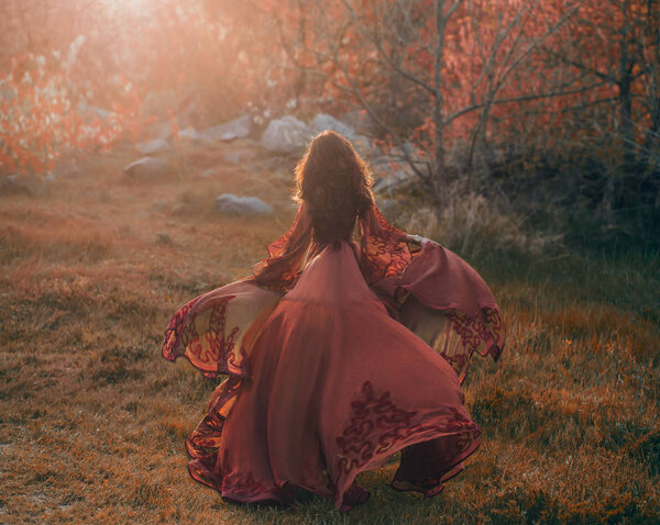 A brunette girl with wavy, thick hair runs to the meeting of the sun. Photo from the back, without a face. The princess has a luxurious, red dress that flutters in the wind. Background sunset