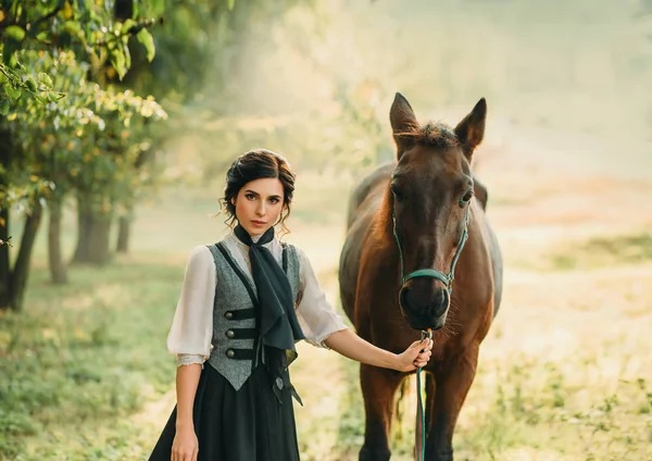 A young lady in a vintage dress strolls through the forest with her horse. The girl has a white blouse, a jabot, a tie, a gray vest, a black long skirt with a train. An ancient, collected hairstyle.
