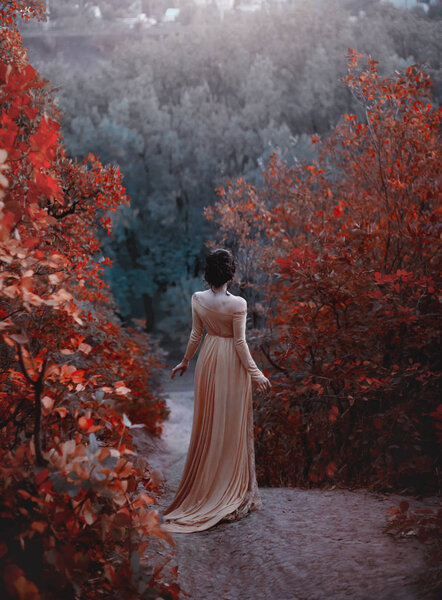 The princess in a yellow vintage dress in the Renaissance is walking along the picturesque autumn hills at dusk. Photo of a brunette girl with a back without a face. Artistic Photography