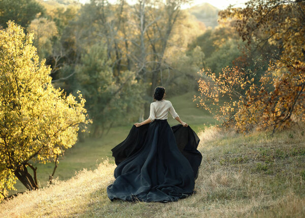 Princess in a vintage dress. Walk along the picturesque autumn hills at sunset. A long train of black skirt fluttered on the run. Photo of a brunette girl with a back without a face. Art Photo