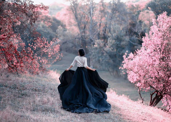 Princess in a vintage dress escapes. Walk through the picturesque autumn hills at sunset in pink tones. A long train of skirt fluttered on the run. Photo of a brunette girl with a back without a face
