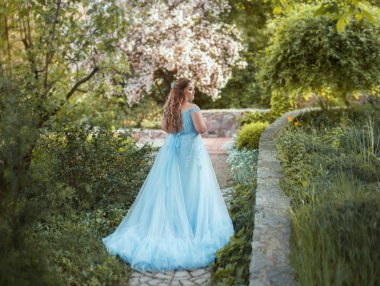 Büyük, güzel, Genç kadın uzun bir tren bir lüks mavi elbiseli bir çiçek bahçesinde yürüyor. Model boyutu artı. Saç kıvırcık ile büyüktür. Sanatsal fotoğraf.