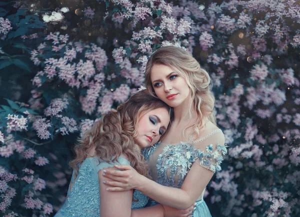 Beautiful big woman hugs a fragile blonde girl. Two princesses in luxurious blue dresses against the background of flowering trees. The model size is plus. Artistic photo.