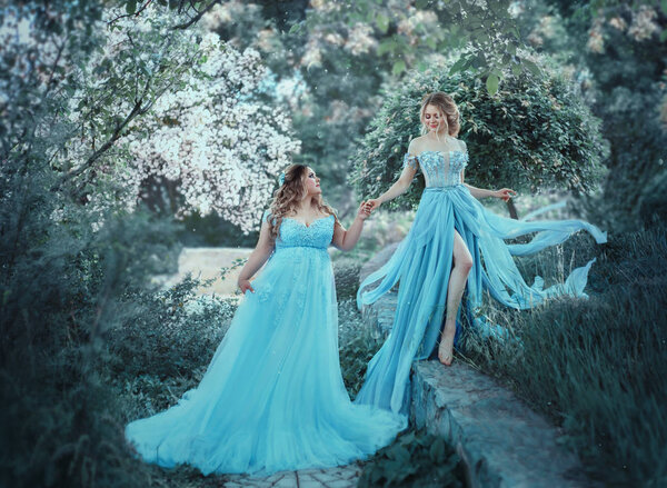 A beautiful big woman is holding a fragile blonde girl in her hand. Two princesses in luxurious blue dresses against the background of flowering trees. The model size is plus. Artistic photo.