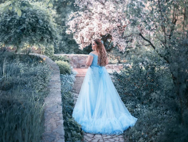 一个高大 年轻的女人穿着豪华的蓝色连衣裙 长着一列火车正在盛开的花园里散步 模型大小为加号 头发很大 有卷发 艺术照片 — 图库照片