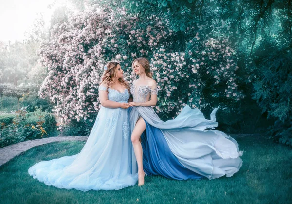 年轻的 成年的女人拥抱在一个开花树的背景 一个神话般的花园 公主穿着漂亮的灰蓝色连衣裙 有一辆长火车在风中摇曳 美妙的修饰 — 图库照片