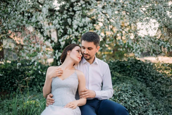 A loving couple hugs in a blooming garden. Red-haired girl in a modest, gray dress in rustic style. Stylish and discreet wedding day. A neat, collected hairdo in an airy beam. Art photo.