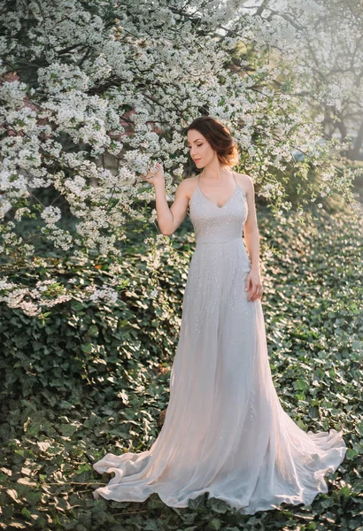 Red-haired girl in a modest, very long, gray dress in rustic style. Portrait of the bride against the background of a flowering tree. A neat, collected hairdo in an airy beam. Glare of the sun.