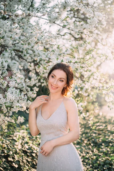 Red-haired girl in a modest, gray dress in rustic style. Portrait of the bride on the background of a blossoming tree with a minimal retouch. A neat, collected hairdo in an airy beam. Nude makeup