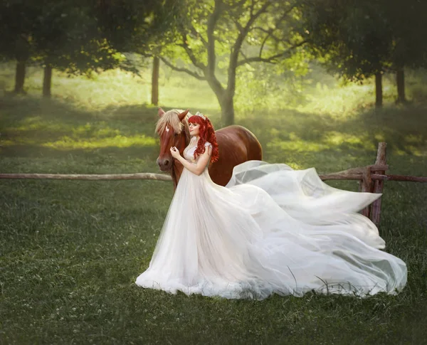 一个年轻的 红头发的公主在水晶头饰 拥抱和抚摸她的马 背景是一个梦幻般的森林空地和围栏 在皇后是一个豪华的 飞行礼服与长的火车 — 图库照片