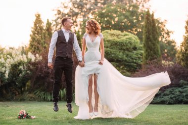 bir atlama içinde neşeli yeni evliler gülmek ve fly air ellerini bir arada yeşil bir bahçe içinde tutan. Gelin uzun, lüks beyaz gelinlik onun sevgili damat görünüyor. sanat fotoğraf.