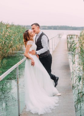 mutlu genç çift, bir uzun ışık beyaz muhteşem gelinlik ve karısı, sarışın bir gelin bekliyoruz köprüde, adam yavaşça kız kucaklayan, yanağını öper. romantik sanat fotoğraf.