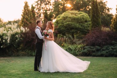 Genç adam bir beyaz uzun gelinlik, şirin bir buket pembe çiçek onun geri, yavaşça çenesinin yüzünü yakın tutarak arkasında tutarak sevgilisini şefkatle kucaklayan. romantik sanat fotoğraf.