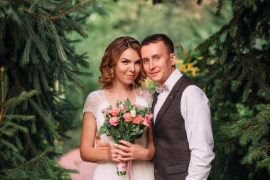 mutlu genç çift, bir sarışın gelin buket pembe çiçek ve damat, yavaşça onun kız kucaklayan tutarak hafif beyaz muhteşem gelinlik, çam ağaçları, güleryüzlü kameraya yakın duran.