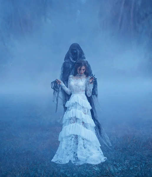 黑魔王的新娘在白色复古网眼半透明连衣裙和一条银色项链站在冰冻的草地上 在一个厚实的蓝色雾在怪物手中 万圣节服装的想法 艺术照片 — 图库照片
