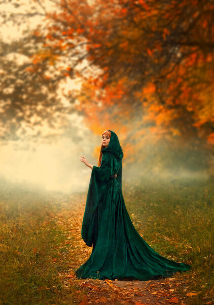 Mysterious stranger the girl turned around on a path in the forest, in a green emerald dress with a hood and wide sleeves, stands in the oanzhevom autumn forest, a sorceress with fiery red hair.