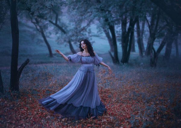 A mysterious girl with wavy dark hair is dancing alone on fallen autumn leaves in a gloomy night forest in a long wonderful blue vintage dress, fabulous heroine, knows no ills, the legend of Pandora.