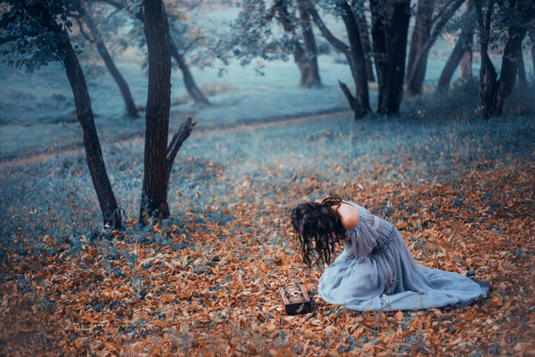 Dark-haired girl in a long, light gray, old-fashioned dress, sits on cold ground covered with autumnal fallen leaves, in a dark forest, crying over Pandora's open box. no face on art photo, depersia.