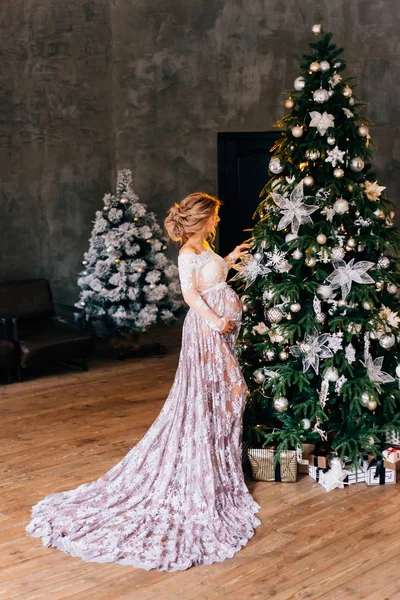 Linda mulher grávida com loira incrível cabelo puro, vestindo um vestido de renda branca translúcida luz com uma tonalidade roxa, decora uma árvore de Natal verde, preparando presentes para o ano novo, sem rosto — Fotografia de Stock