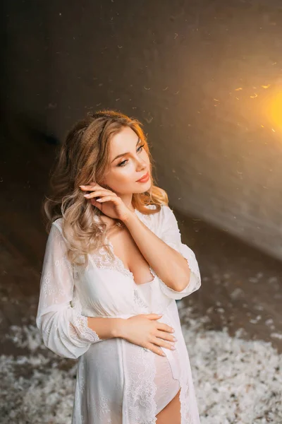 Cute tender attractive pregnant girl dressed in a wonderful white light nightie, touches the hair with her hand, hugs her tummy, neat makeup on her face with a soft light, surrounded by feathers — Stock Photo, Image