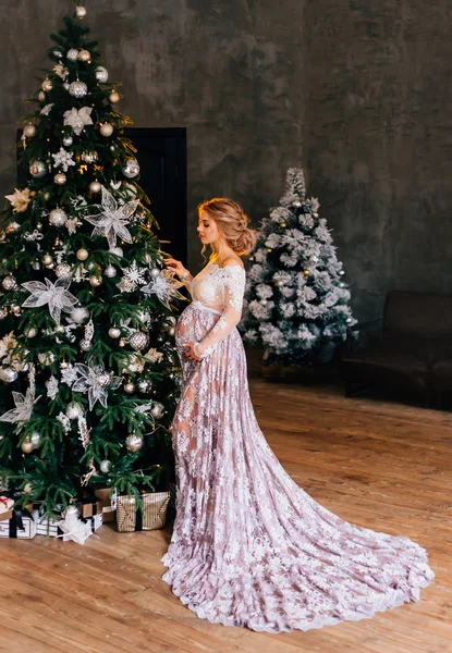 Mulher grávida bonita com cabelo encaracolado incrível loiro puro, vestindo um vestido leve translúcido rendas brancas com uma tonalidade roxa, decora a árvore de Natal, preparando presentes de ano novo, esperando por um milagre — Fotografia de Stock