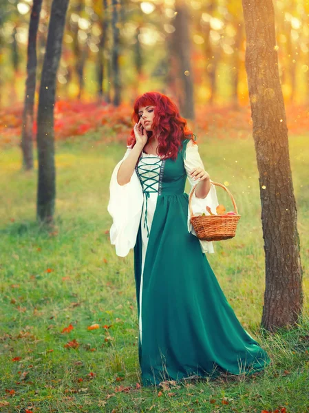 Girl with fiery red hair, an unusual appearance, in an emerald white gorgeous delightful dress, holds a basket and explores new paths of the forest, a bright photo in the glare of the morning sun — Stock Photo, Image