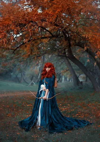 Загадочная рыжеволосая воительница стоит на страже своей земли, эльфийская принцесса держит лук и стрелы, готовясь к бою, привлекательная женщина в туманном лесу одна, готический стиль, прохладные цвета — стоковое фото
