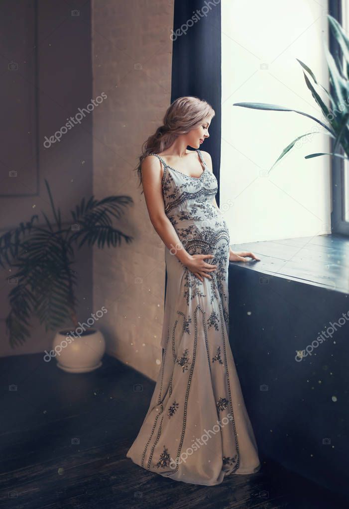 mysterious pregnant queen, woman stands by the window in loft room, the sunlight illuminates, blond curly hair gathered into tail, dressed in gray long decorated dress, holds her tummy with one hand