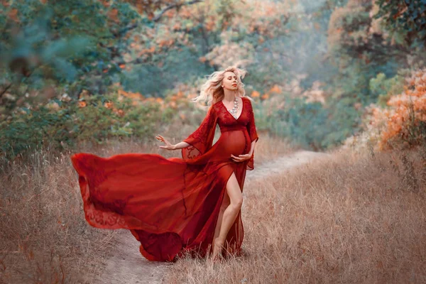 Jovem mulher grávida com cabelo encaracolado loiro em luz longa batendo vestido vermelho escarlate com pernas expostas nuas e colar brilhante está de pé na floresta, segurando, abraçando sua barriga. arte outono foto — Fotografia de Stock