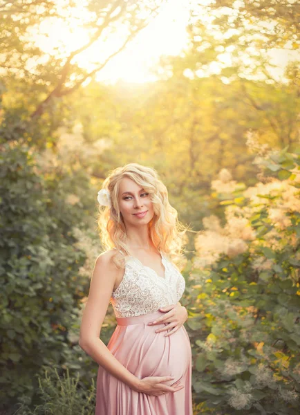 Amable hermosa mujer embarazada joven está sonriendo, usando un vestido elegante rosa con encaje top blanco en los suaves rayos del sol con un maquillaje suave. pelo rizado claro decorado con orquídea . — Foto de Stock
