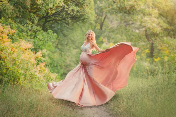 Ein süßes frühlingsschwangeres Mädchen mit leicht gelocktem Haar tanzt im Wald, trägt ein zartes langes, elegantes, fließendes rosa Kleid mit weißem Spitzenoberteil, lächelt in die Kamera und umarmt ihren Bauch. Kunst — Stockfoto