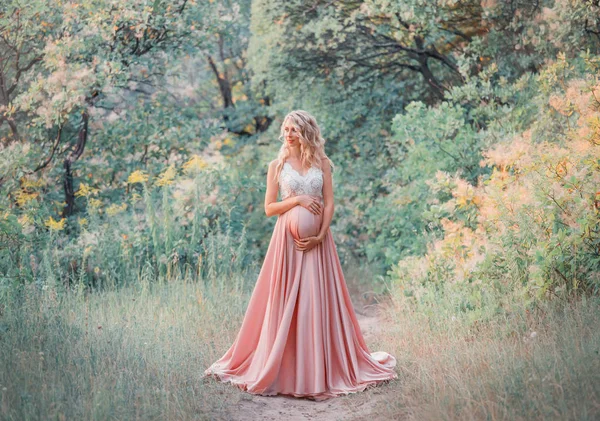 Молодая милая беременная девушка со светлыми вьющимися волосами обнимает и держит животик, стоя в волшебном лесу в холодных оттенках. одевается в длинное элегантное атласное шелковое розовое платье с кружевным верхом. обработка фотографий — стоковое фото