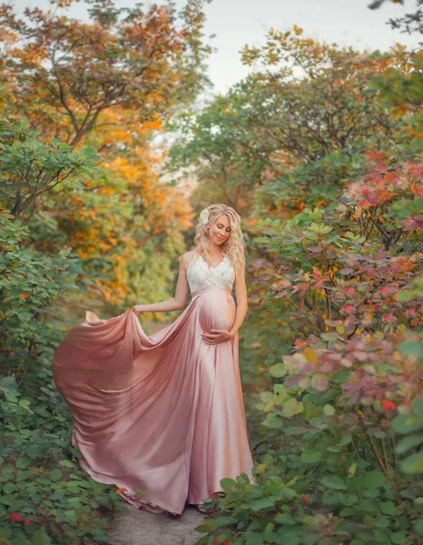 Mladá těhotná žena s blond vlasy s živou orchideje je oblečená v dlouhé růžové luxusní vlající šaty s bílý krajkový top, stojí na cestě v lese a úsměvy, objetí její bříško — Stock fotografie