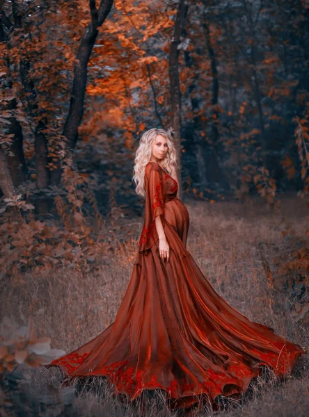 Jovem grávida com cabelo encaracolado loiro em luz longa batendo vestido vermelho escarlate, de pé na floresta, segurando, abraçando sua barriga. como uma fada magnífica rainha. arte outono escuro halloween foto — Fotografia de Stock