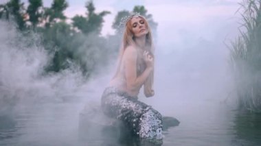 bir kuyruk ile gerçek deniz kızı kayaların üzerinde oturur ve sis sarışın saç örgü