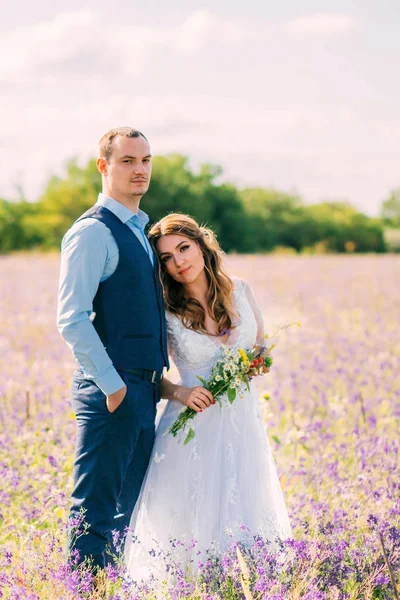 Merveilleux jeune couple, mariée repose sur la poitrine de son marié bien-aimé, se tenir dans un champ avec des fleurs violettes, fille dans une robe élégante blanche, tient des fleurs sauvages, l'homme dans un costume de mariage bleu et chemise — Photo