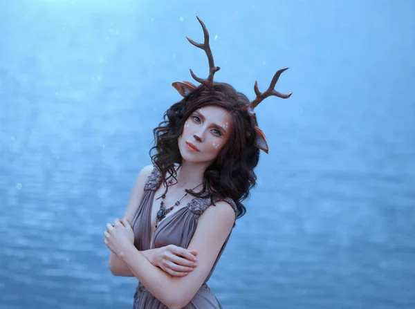 Chica en la imagen de un faun, traje y maquillaje de un ciervo, un personaje fantástico del espíritu del bosque en vestido marrón, foto de retrato de una dama triste en el fondo del lago mágico, tonos frescos azules —  Fotos de Stock