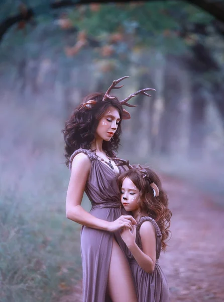 Казкова мати оленя на шляху обіймає дитину і тримає руку, закриває очі і слухає звук туманного лісу, в довгих коричневих сукнях з фіолетовим відтінком, виріз показує ногу жінки — стокове фото