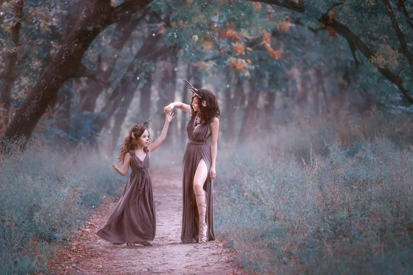 Splendida donna in costume da cervo sta girando la figlia su un sentiero nel bosco, indossando abiti lunghi marroni, mostrando la sua gamba affascinante in un taglio, favolosi padri si divertono e ballano, tiro creativo — Foto Stock