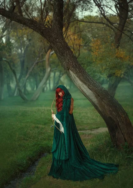 Cazador, chica pelirroja con un lazo en las manos en busca de la víctima, vestida con terciopelo verde esmeralda y un impermeable, mirando directamente a la cámara, caminando sola en el bosque lleno de niebla — Foto de Stock