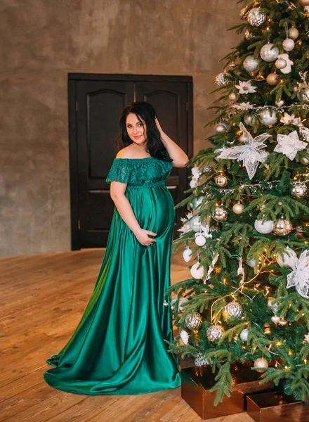 Hermosa chica encantadora en vestido largo y encantador verde esmeralda en la habitación espaciosa cerca del árbol de Navidad decorado, endereza su peinado de pelo oscuro, sonríe dulcemente, abrazando suavemente la panza —  Fotos de Stock