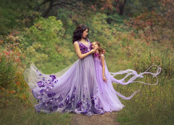 Foto di famiglia di mamma e figlia in lussuosi abiti viola svolazzante con fiori, sono in piedi nella foresta estiva. Carina signora abbraccia la ragazza, sorride, guardandola. foto d'arte in colori caldi — Foto Stock