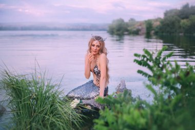 rüya gibi sarışın perisi deniz diadem ile yalnız nehre oturur ve aşağı görünüyor sürpriz kaldırdı, elini ağzına bir gümüş ile rüya gibi bir Bayan kuyruk mavi gökyüzü ve parlak yeşil çim karşı